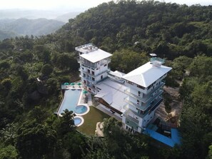 A multi-story hotel with a swimming pool and a tennis court, surrounded by lush greenery.