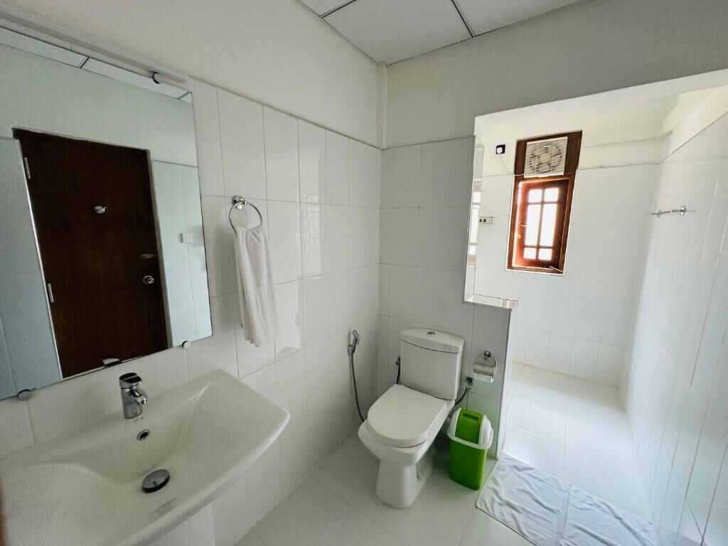 A small, clean bathroom with white tiles, a white sink, a toilet, and a green trash bin.