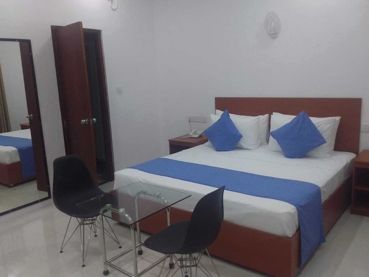 Una habitación de hotel con una cama, dos sillas negras, una mesita de cristal pequeña y una mesita de noche de madera.
