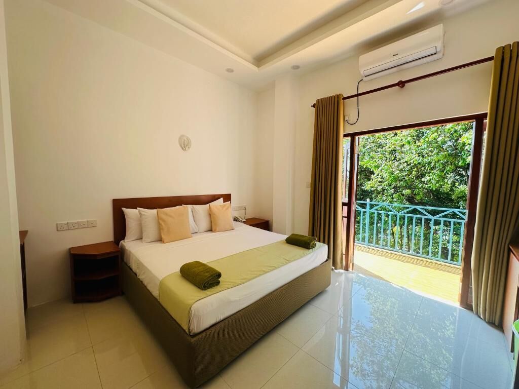 A hotel room with a large bed, a nightstand, a window with curtains, and a view of greenery outside.