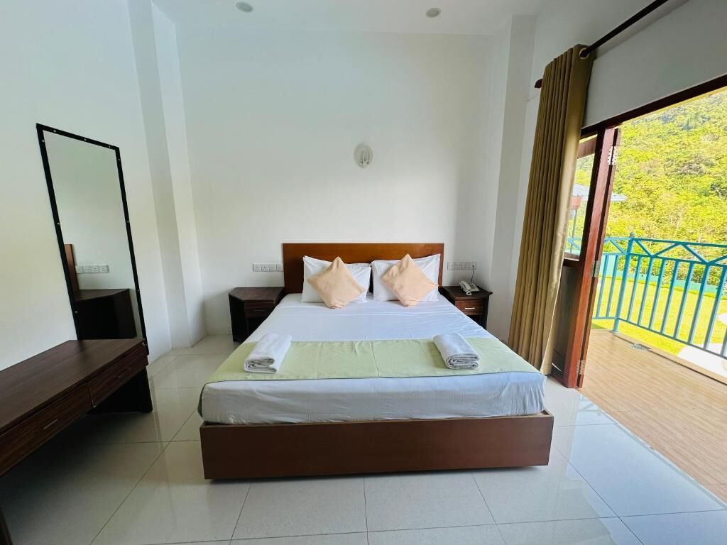 A hotel room with a large bed, a bedside table, a nightstand, a mirror, and a balcony with a view of greenery.