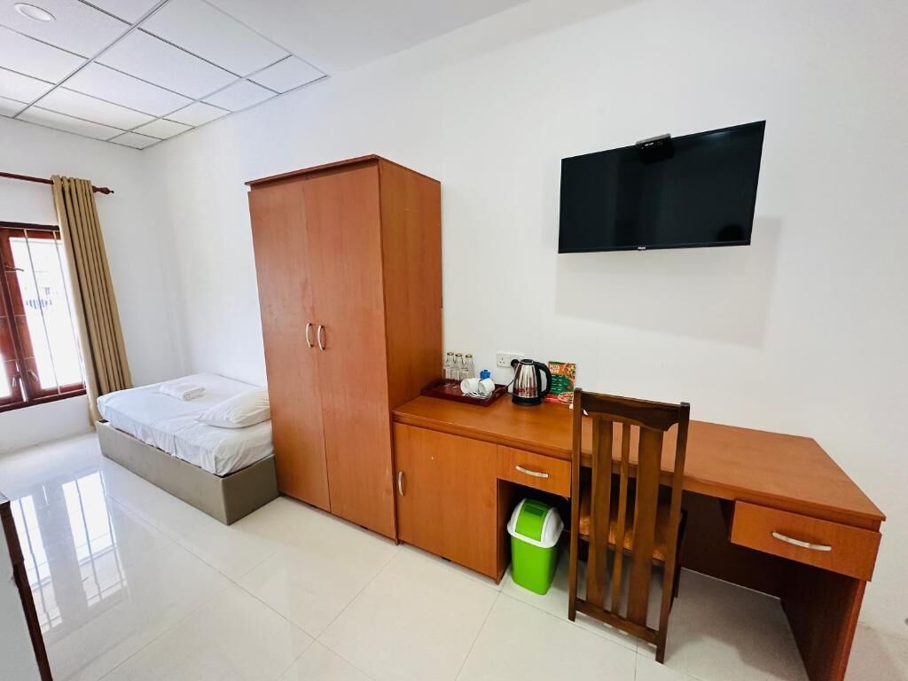 Une chambre à coucher comprenant un lit, une armoire en bois, un bureau, une chaise, un téléviseur et une poubelle verte.