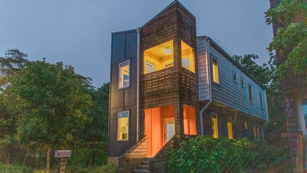 A modern two-story house with a dark exterior, large windows, and a staircase leading to the entrance.
