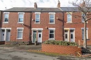 A row of red brick terraced houses with white-framed windows and doors, a small garden area with a tree, and a clear sky.
