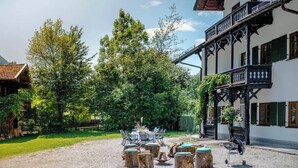 An outdoor seating area with round wooden tables and chairs, surrounded by trees and a building with balconies. - Cozy mountain loft - 15 minutes from Garmisch (Oberau)