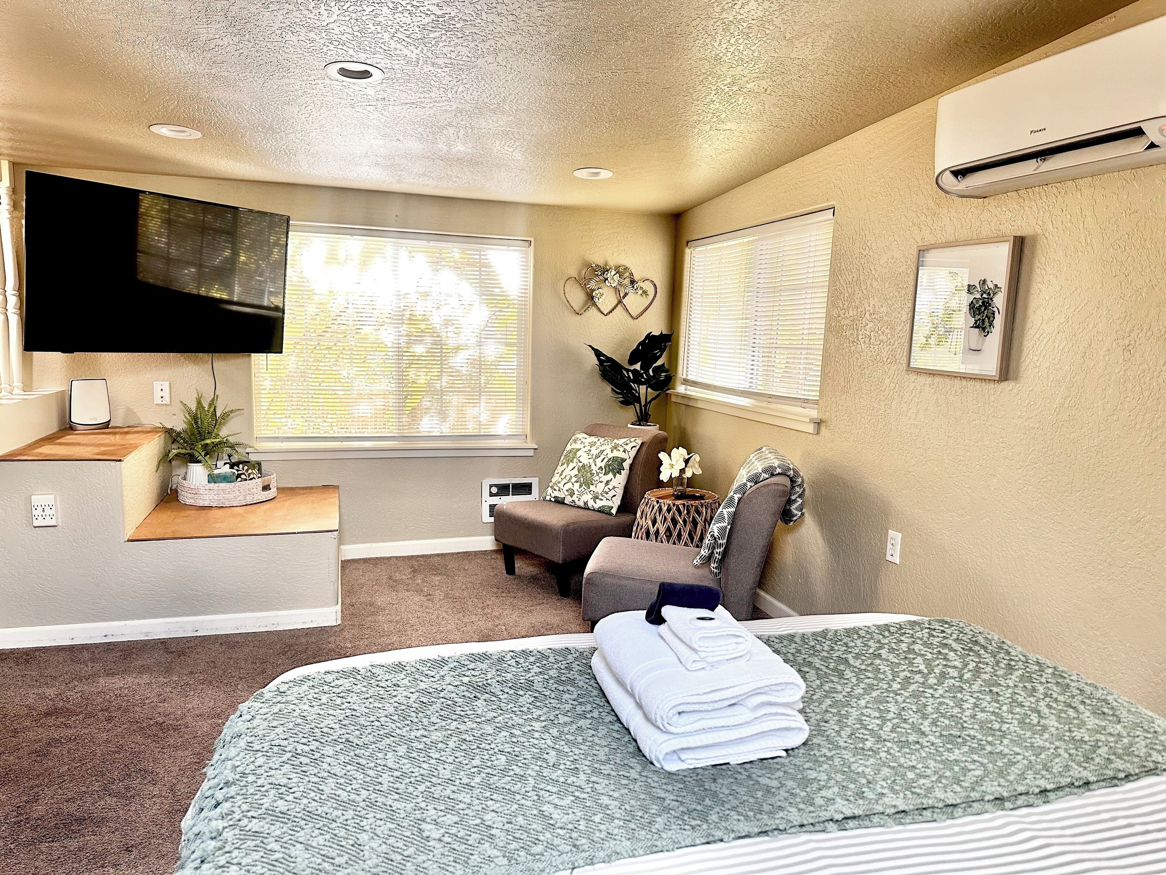 A bedroom with a bed, a TV, a desk with a computer, a window with blinds, and a wall-mounted air conditioner.
