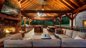 An outdoor covered patio with stone walls, a stone fireplace, wooden furniture, and a view of the surrounding forest.
