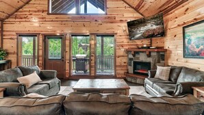 A log cabin interior with a fireplace, leather sofas, a coffee table, and large windows.