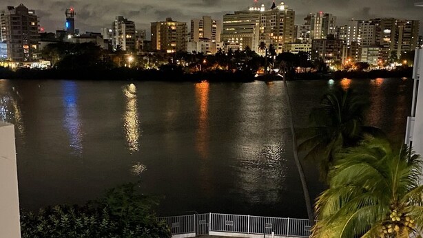 A nighttime view of a waterfront property with a swimming pool, palm trees, and a city skyline in the background. - Beautiful apartment at the heart of Condado Beach (San Juan)