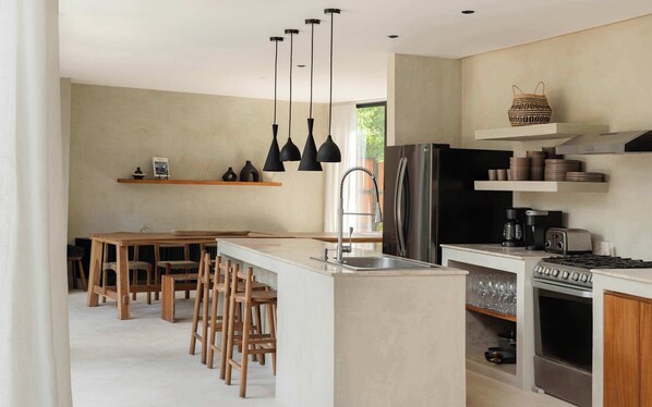 A modern kitchen with a central island, bar stools, a stainless steel refrigerator, a built-in oven, and pendant lights. - Palacio Papaya • 2 Pools, Rooftop Bar & Pickleball (Tulum)