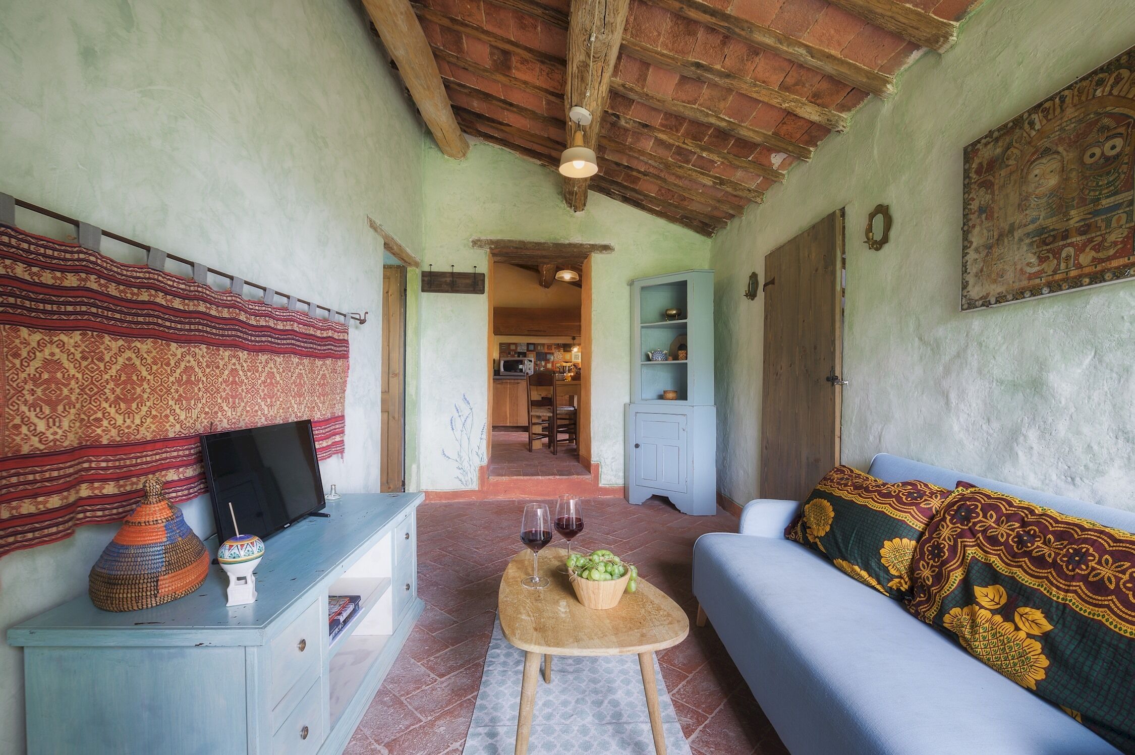 A cozy living room with a sofa, a coffee table with a bowl of fruit and wine glasses, a television on a cabinet, and a patterned tapestry on the wall.
