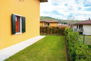 Ein gelbes Gebäude mit Fenster und Tür, ein gepflasterter Weg und ein grüner Hecke.