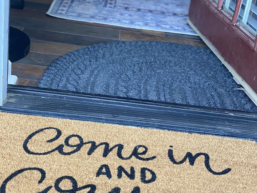 Un felpudo con el texto "Entra y acurrúcate" y una alfombra que conduce a una puerta.