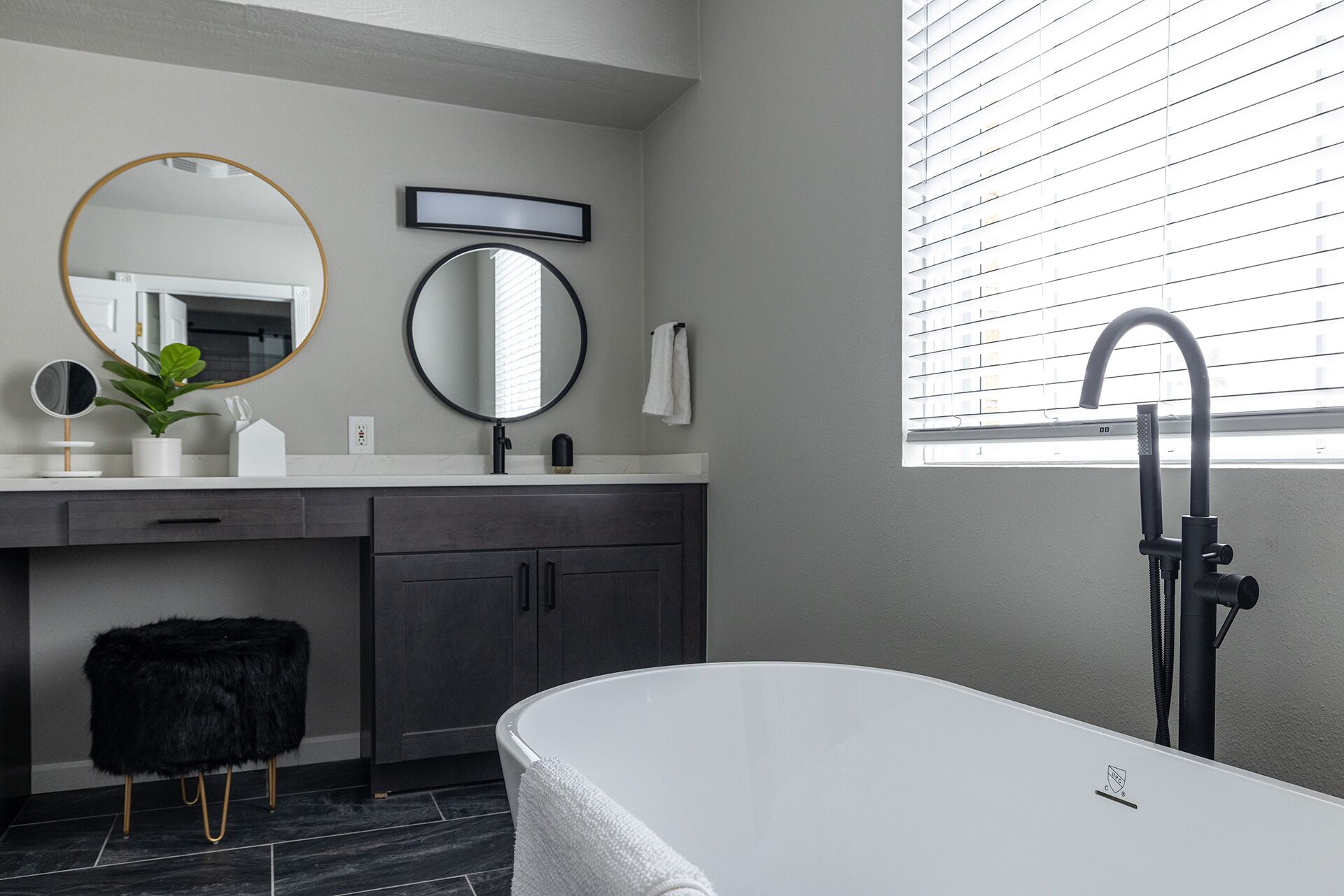 A modern bathroom with a freestanding bathtub, a large mirror, and a window with blinds.