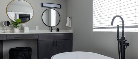 A modern bathroom with a freestanding bathtub, a large mirror, and a window with blinds.