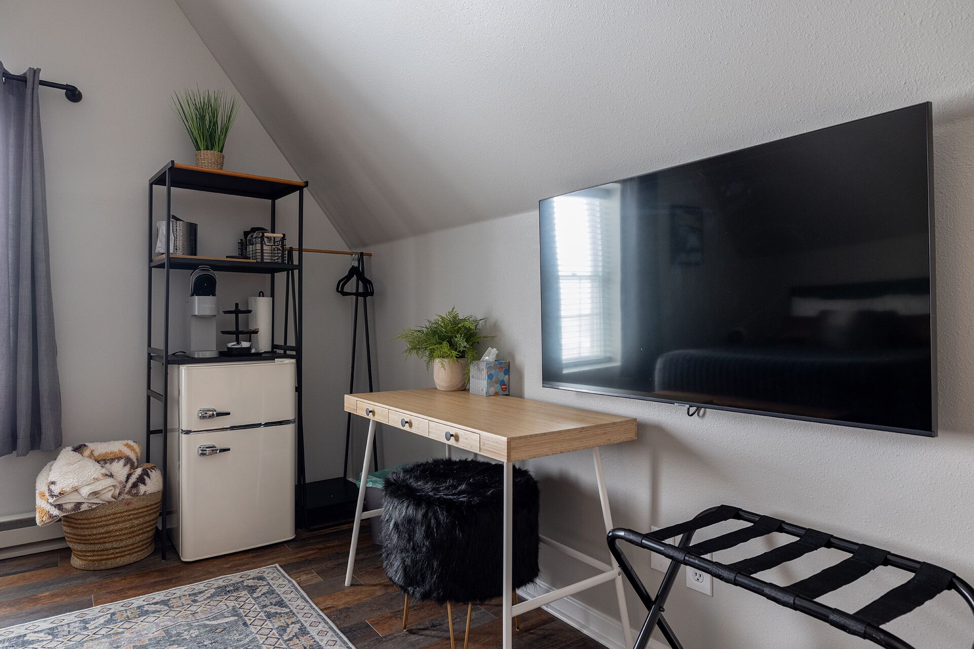 A compact room with a desk, a TV, a small fridge, a plant, and a black stool.