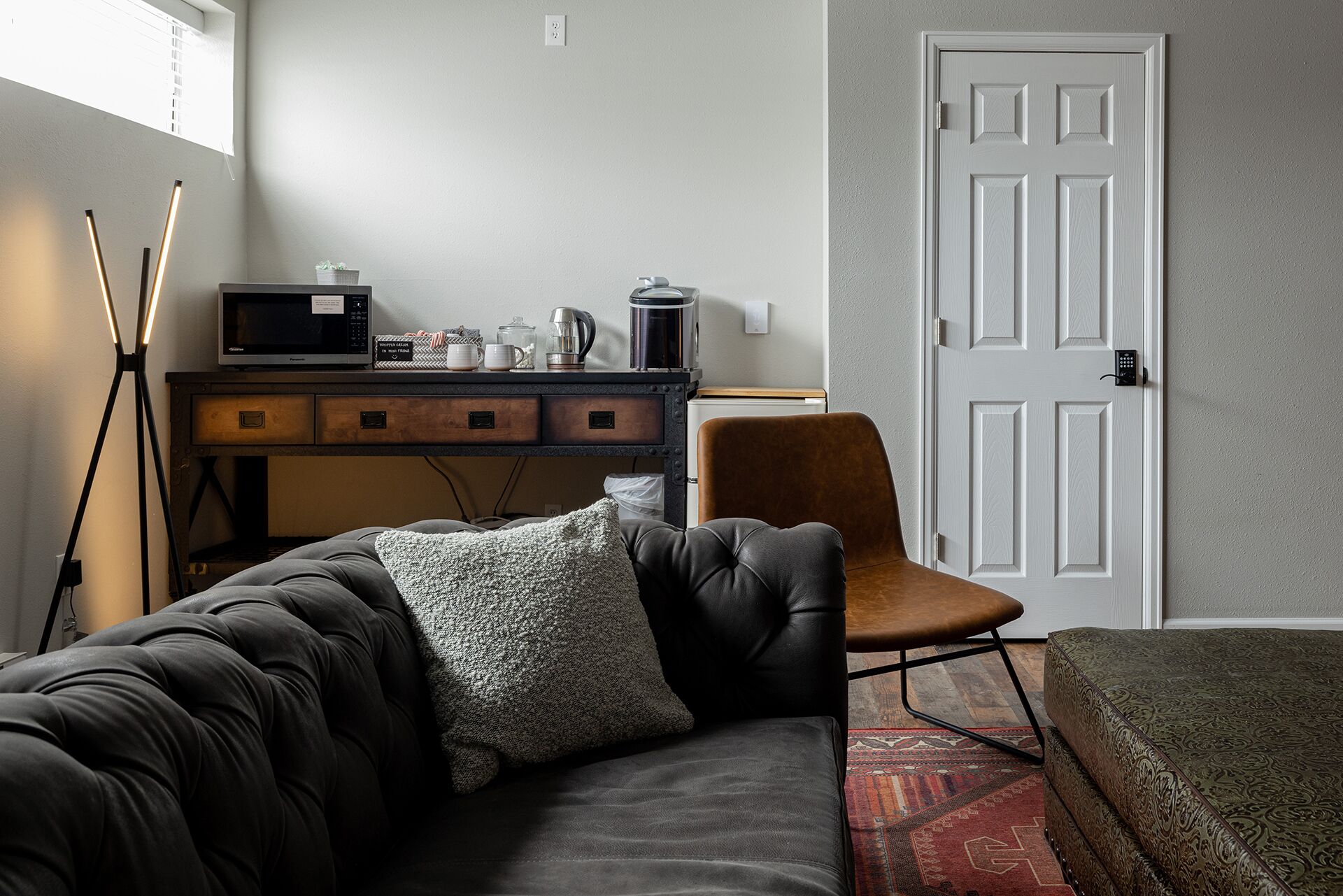 A modern living room with a dark leather sofa, a brown armchair, and a patterned rug. A desk with a microwave and various items is against the wall, and a white door is visible.
