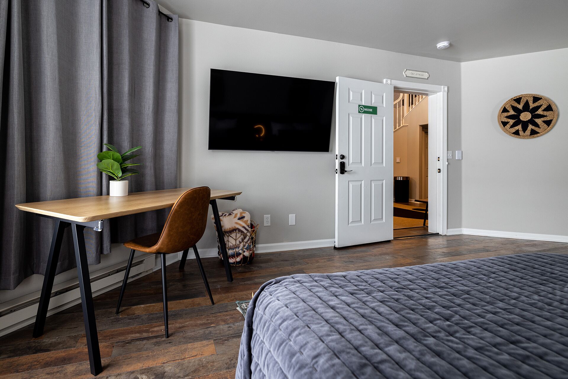 A hotel room with a bed, a desk with a chair, a television, a door with a sign, and a dartboard on the wall.