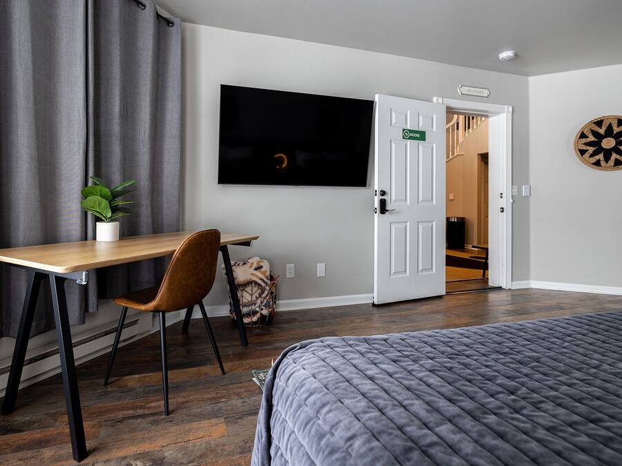 A hotel room with a bed, a desk with a chair, a television, a door with a sign, and a dartboard on the wall.