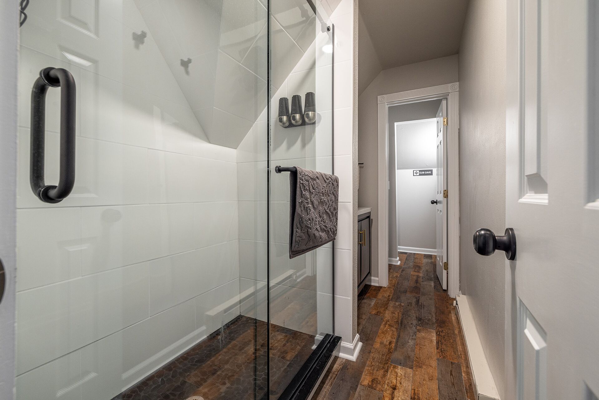 A modern bathroom with white tiles, a glass shower enclosure, a towel rack, and a door leading to another room.