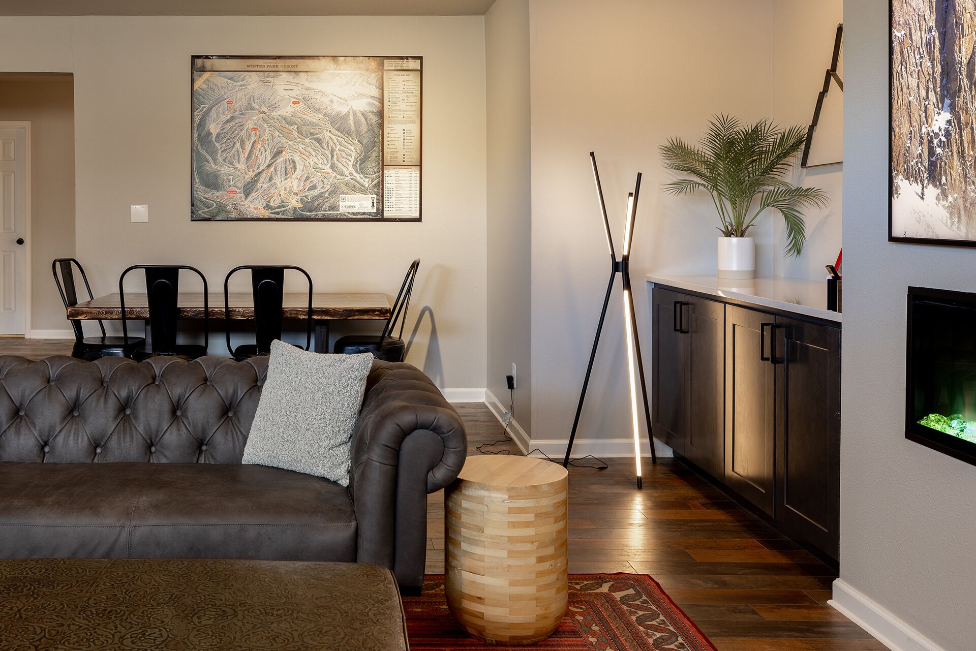 Una sala moderna con un sofá de cuero oscuro, un comedor con mesa y sillas de madera, un cuadro abstracto grande, una lámpara de pie alta y una planta pequeña en la encimera de la cocina.