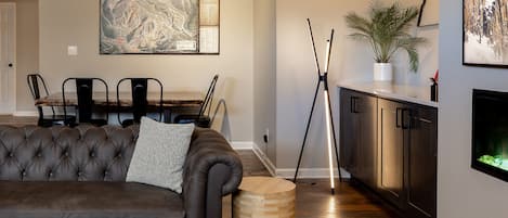 Una sala moderna con un sofá de cuero oscuro, un comedor con mesa y sillas de madera, un cuadro abstracto grande, una lámpara de pie alta y una planta pequeña en la encimera de la cocina.