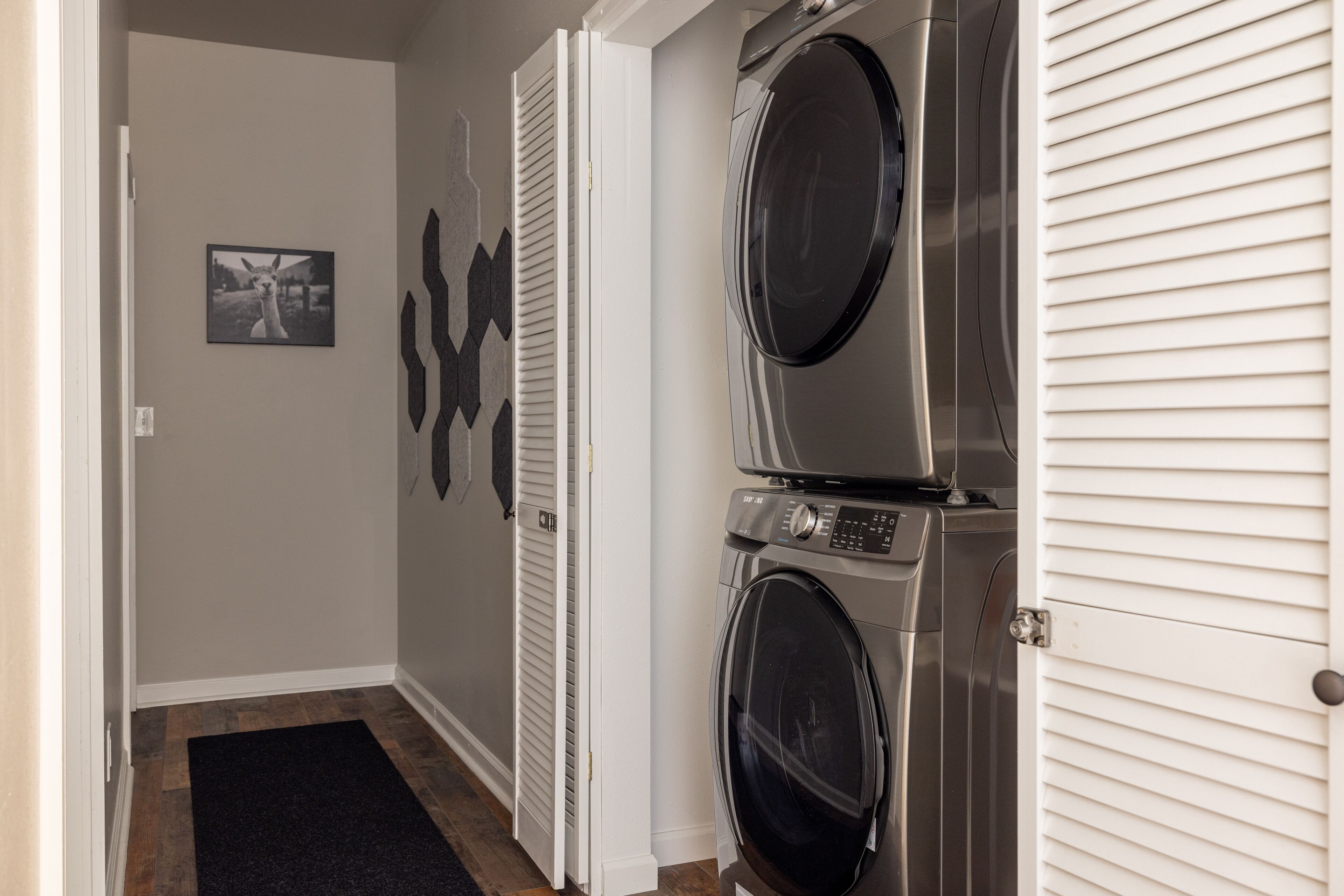 Une buanderie équipée de deux machines à laver et deux sèche-linge à chargement frontal superposés, d’un tapis noir sur le sol et d’un tableau représentant un lama accroché au mur.