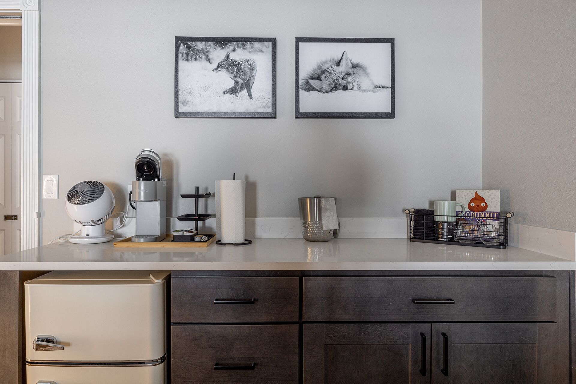 A kitchen with a coffee maker, kettle, and tea kettle on the counter. There are two framed pictures on the wall and a coffee machine on the left.