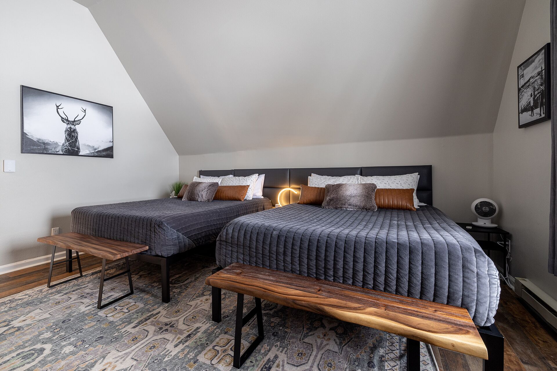 A bedroom with two beds, a bench, a rug, and a picture of a deer on the wall.