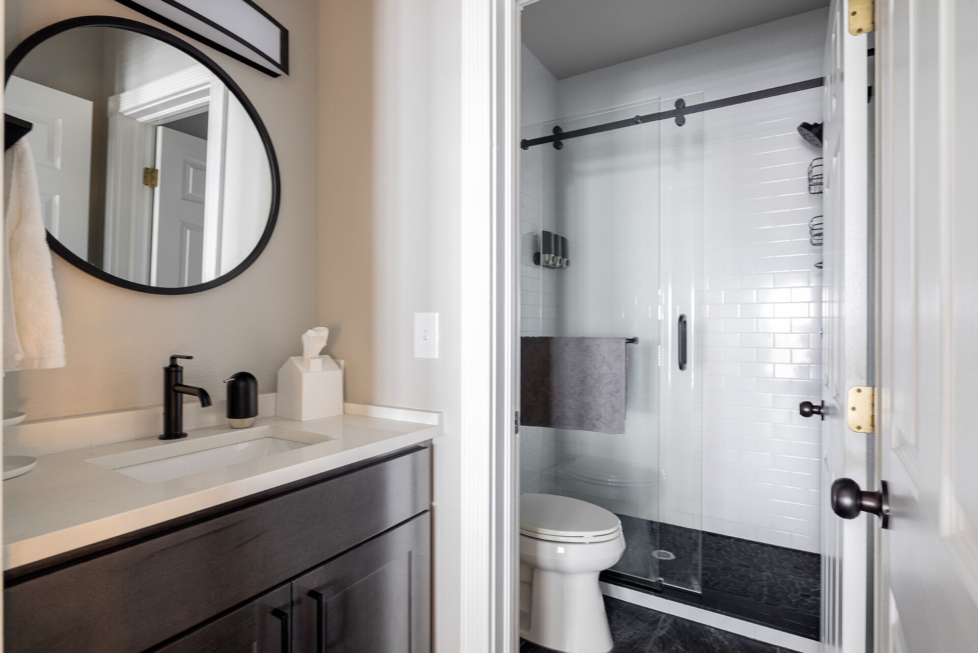 Un baño moderno con un lavamanos blanco, un grifo negro y un espejo encima. Se ve un inodoro y una zona de ducha detrás de una puerta de cristal.