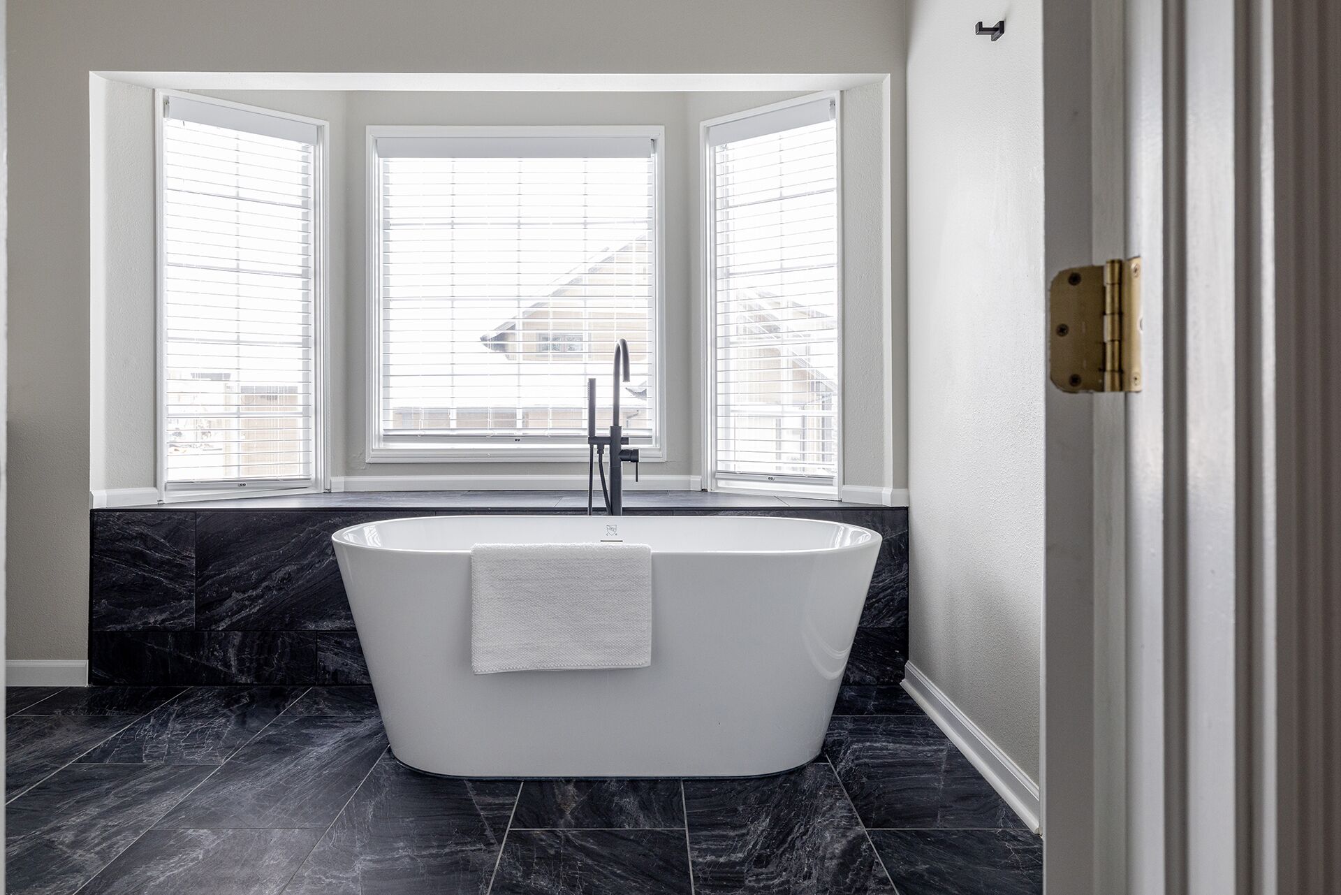A modern bathroom with a freestanding bathtub, large windows, and dark marble flooring.