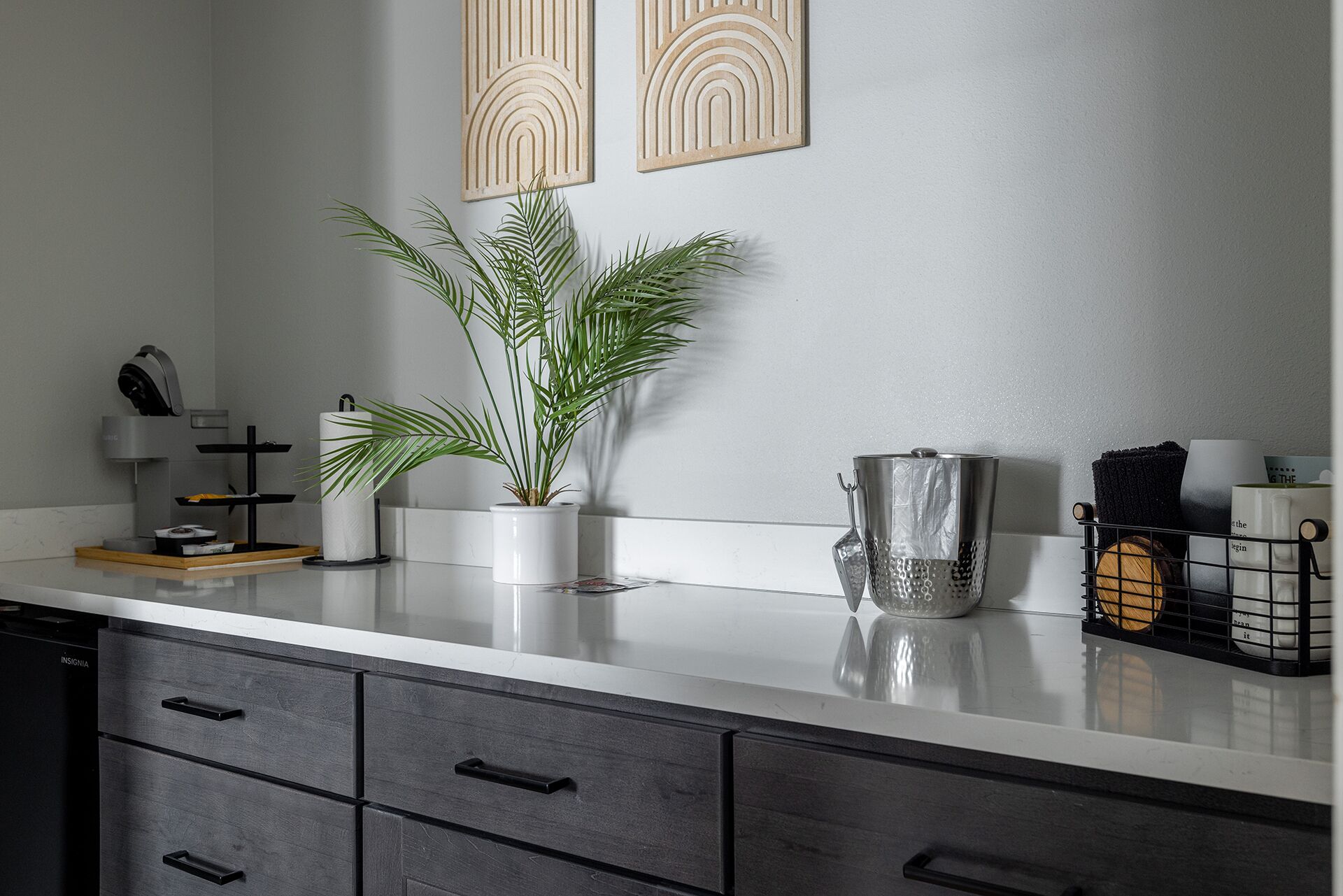Una cocina con encimera blanca, cajones oscuros, una planta en maceta, una jarra de metal y dos paneles decorativos en la pared.