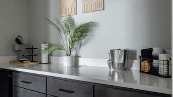 Una cocina con encimera blanca, cajones oscuros, una planta en maceta, una jarra de metal y dos paneles decorativos en la pared.