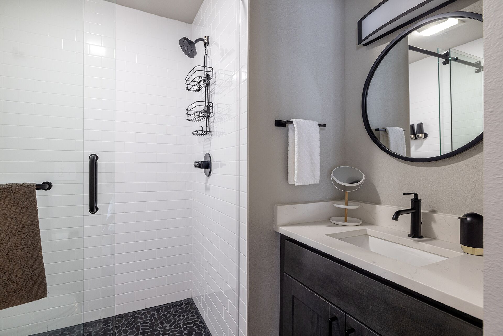 Un baño con paredes de azulejos blancos, una ducha con grifo negro, un tocador con espejo y un perchero para toallas.