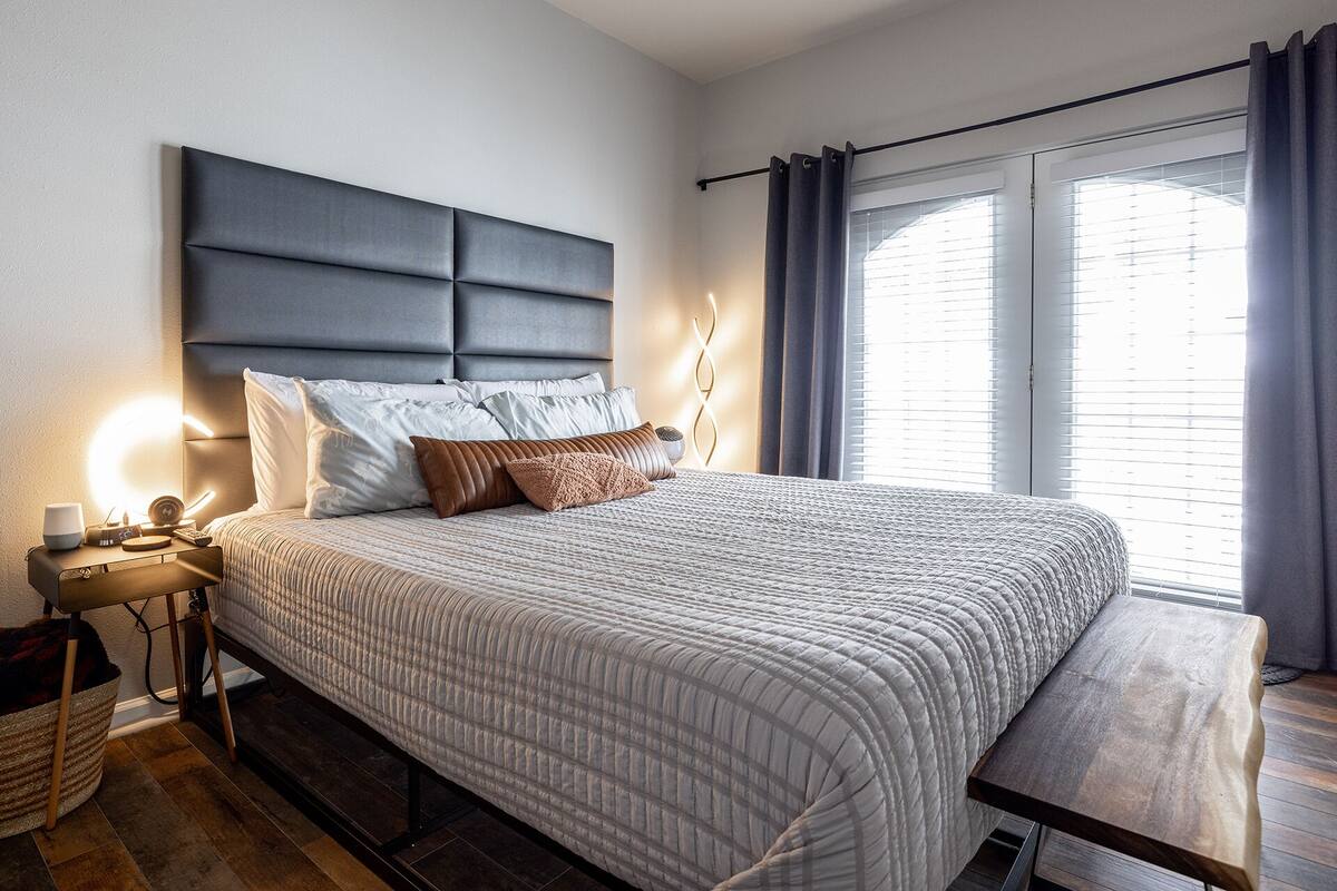 Ein ordentlich eingerichtetes Schlafzimmer mit einem großen Bett, Nachttischen, einer Nachttischlampe und Blick ins Freie durch ein Fenster mit Vorhängen.