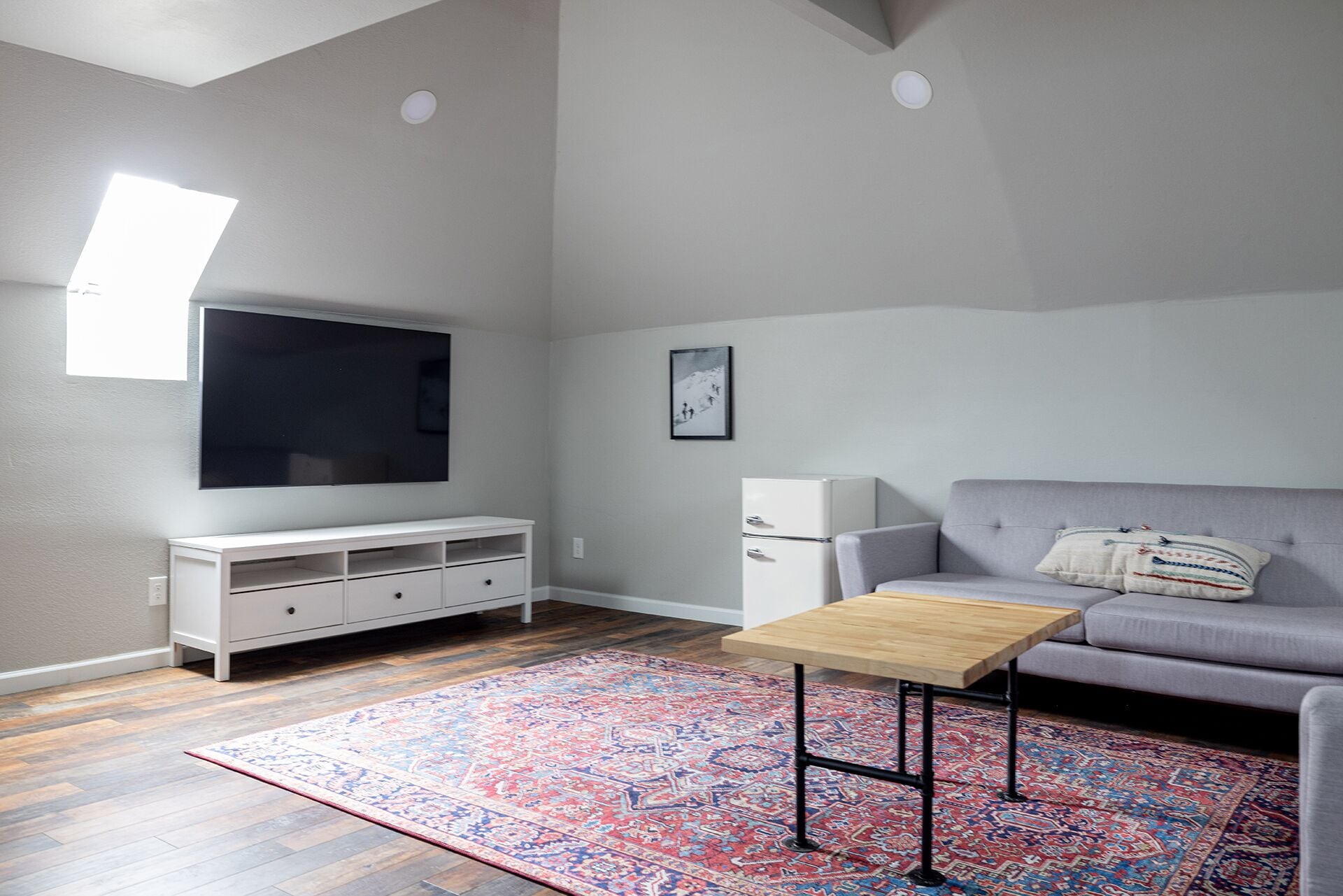 A modern living room with a grey sofa, a wooden coffee table, a white TV stand, and a patterned rug.