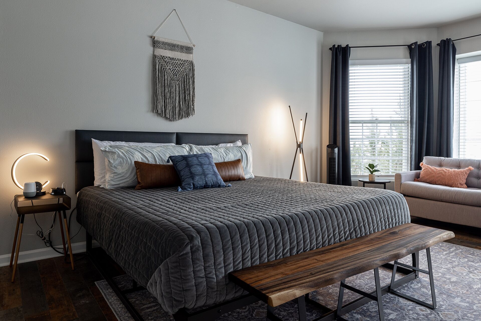 Une chambre à coucher moderne avec un grand lit, des tables de chevet, un banc et une vue sur l’extérieur par une fenêtre.