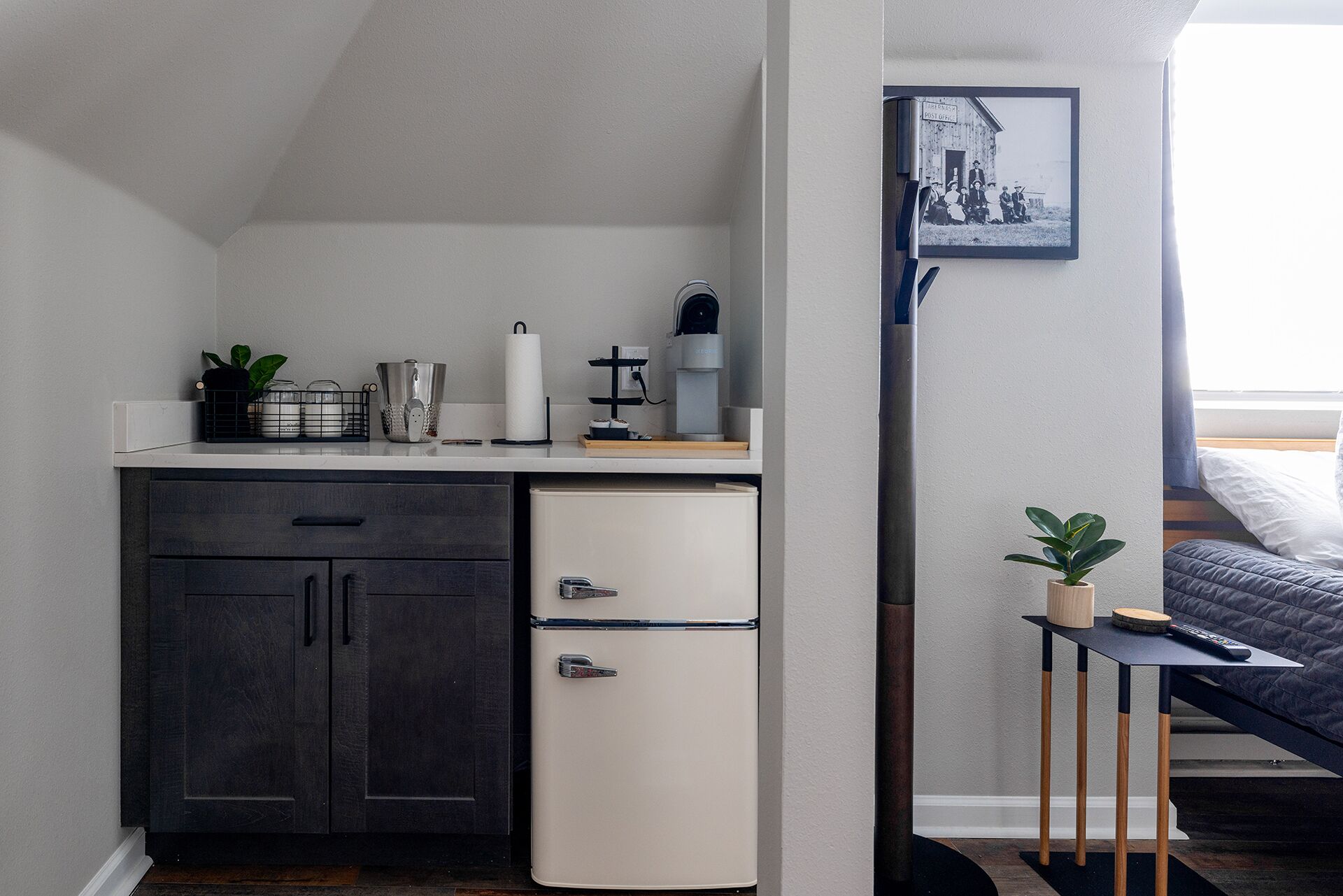 A compact bedroom with a bed, a small bedside table, a desk with a chair, and a refrigerator.