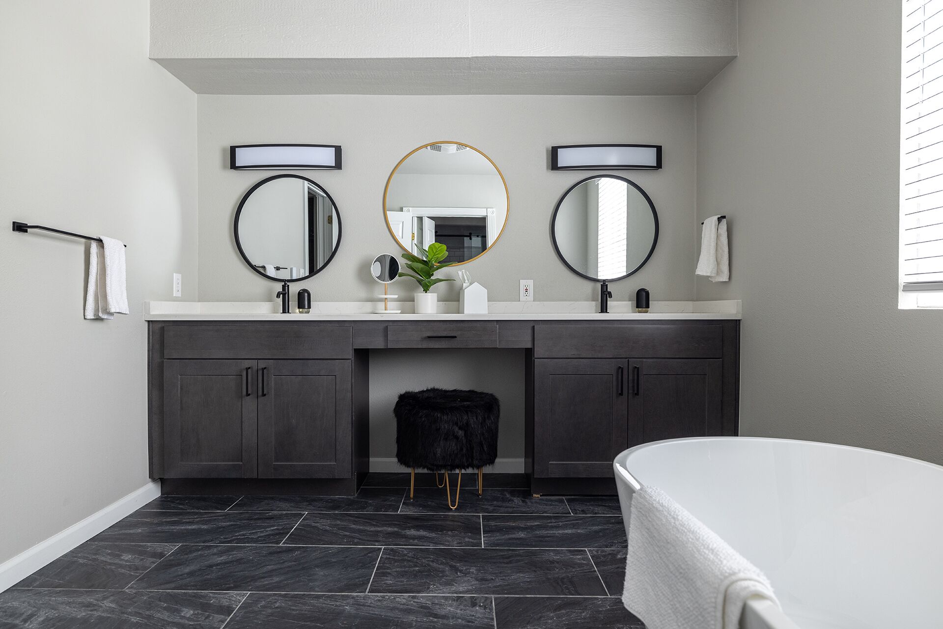 A modern bathroom with a double vanity, two round mirrors, a white bathtub, and dark grey cabinets.