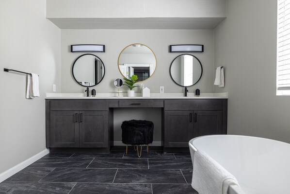 A modern bathroom with a double vanity, two round mirrors, a white bathtub, and dark grey cabinets.