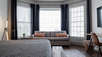 Habitación de hotel moderna con una cama grande, un sofá y un escritorio. Amplias ventanas con cortinas, una lámpara de pie y vistas a la ciudad.