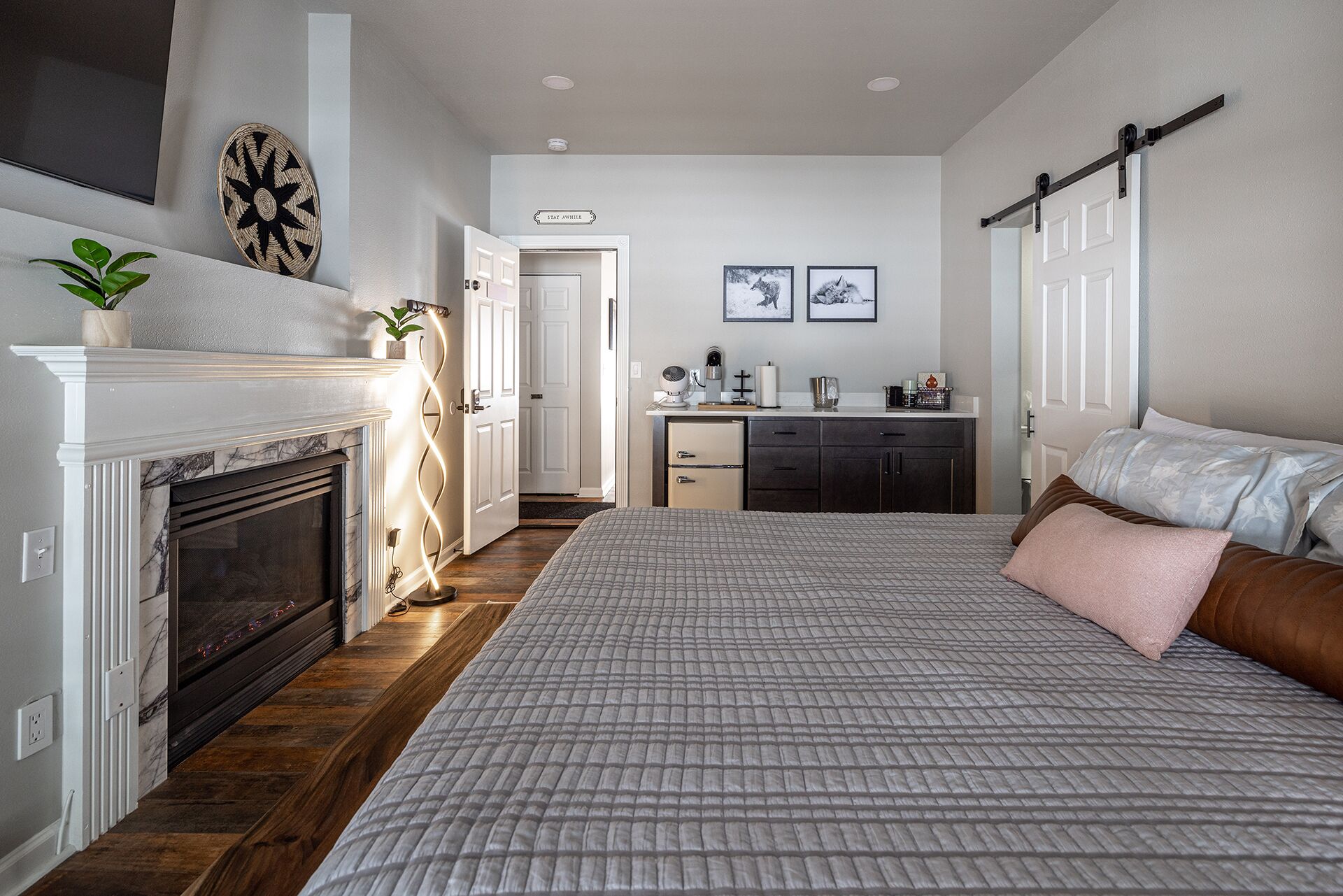A bedroom with a fireplace, a bed with a quilted bedspread, a bedside table, a nightstand, and a small potted plant.