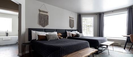 A modern bedroom with two beds, a bench, a hanging light fixture, and a large window.