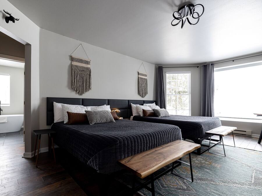 A modern bedroom with two beds, a bench, a hanging light fixture, and a large window.