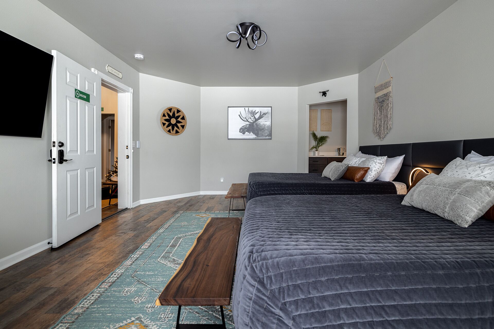 A modern bedroom with a large bed, a bench, a wall-mounted TV, and a door labeled "Moose".