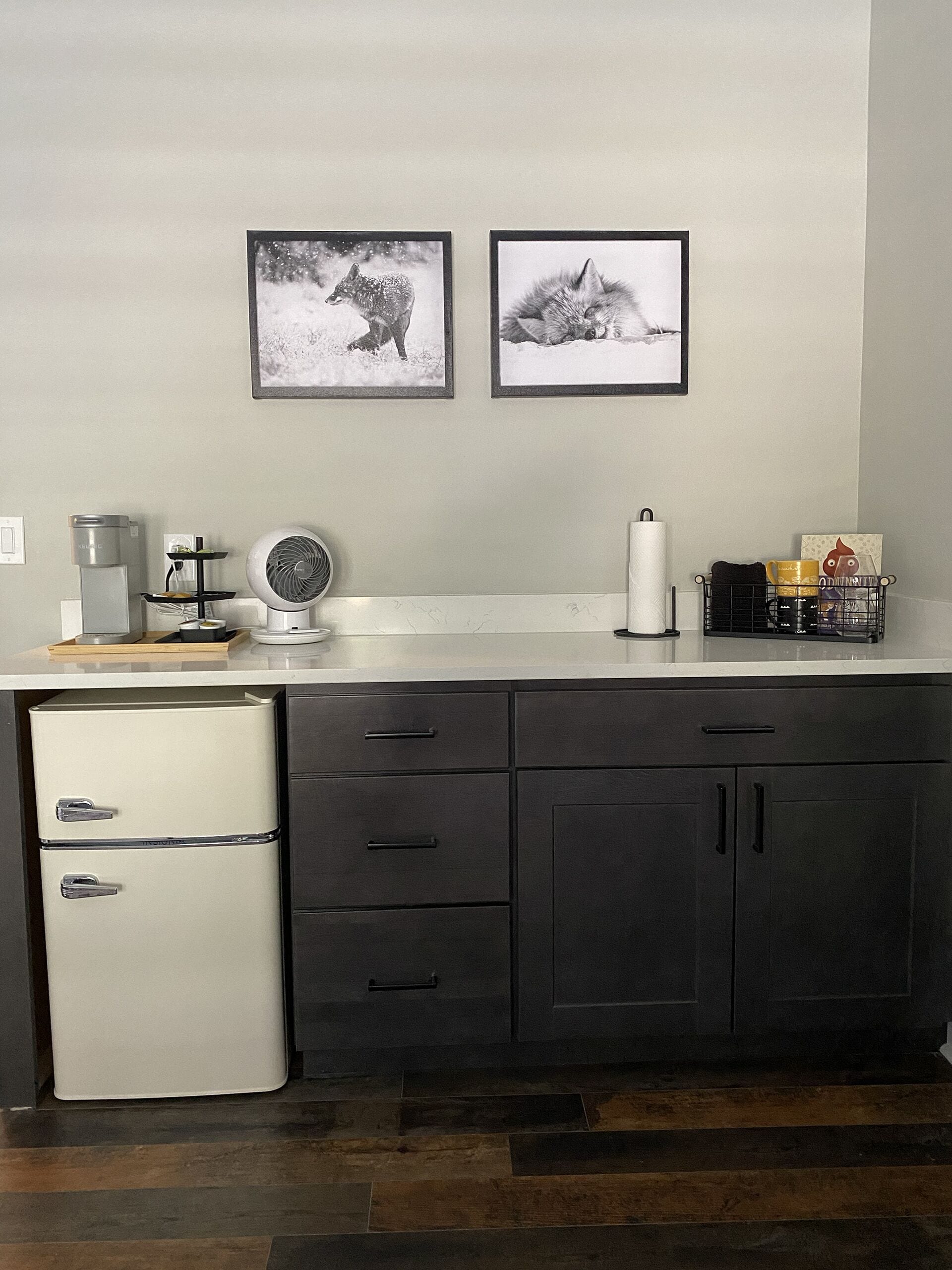 Een keuken met een koelkast, een koffiezetapparaat, een ventilator en twee ingelijste foto's aan de muur.