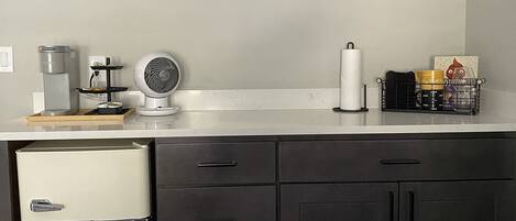 Een keuken met een koelkast, een koffiezetapparaat, een ventilator en twee ingelijste foto's aan de muur.