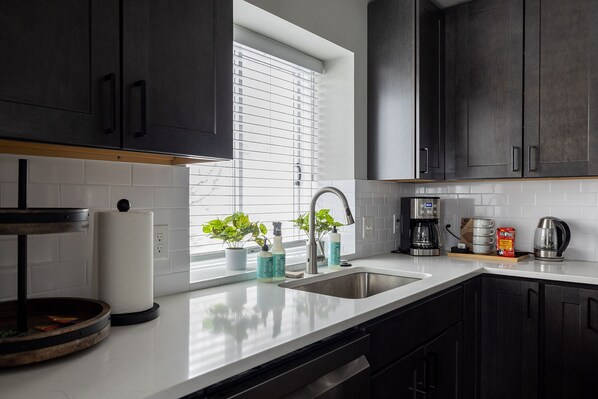 A modern kitchen with dark cabinets, a white countertop, a stainless steel sink, a coffee maker, and a window with blinds.