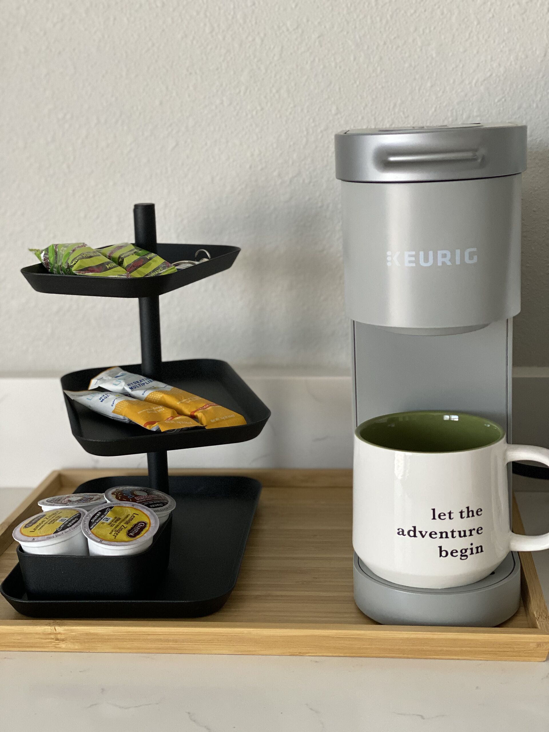 A Keurig coffee maker with a mug that says "let the adventure begin," a tray with food items, and a tray with coffee pods.