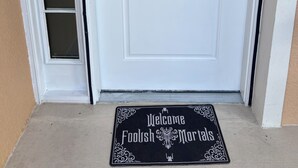 A white door with a wreath and a doormat that reads "Welcome Foolish Mortals".